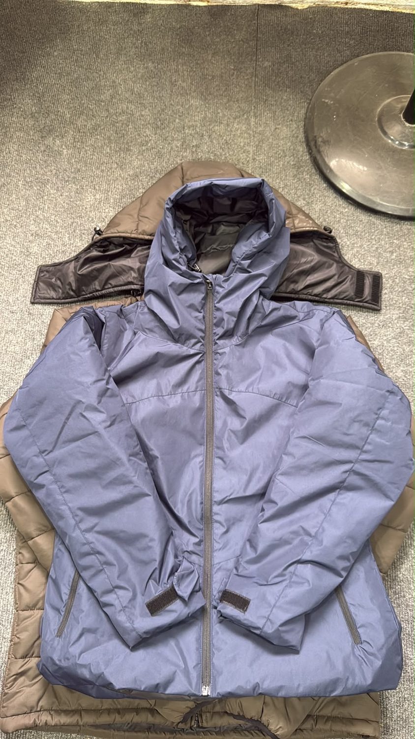 A periwinkle blue jacket laid over a tan jacket on grey carpet with hook and loop tabs at both cuffs
