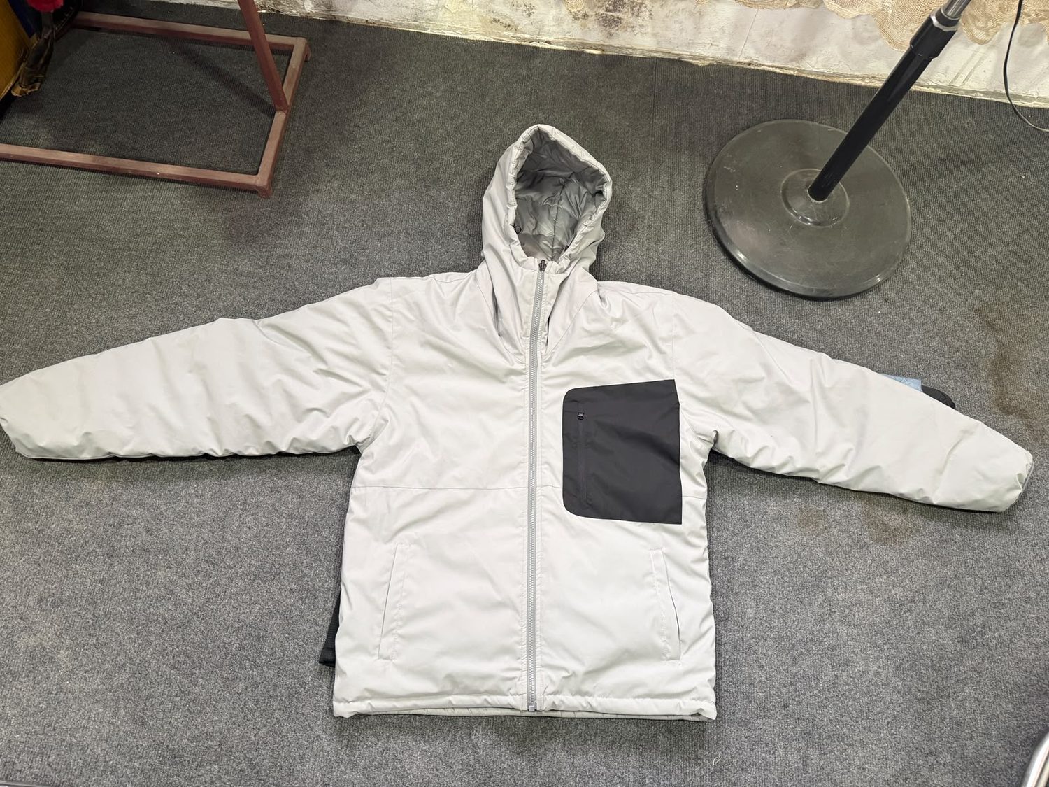 An off white hooded jacket laid flat on grey carpet with a large dark panel pocket on the chest