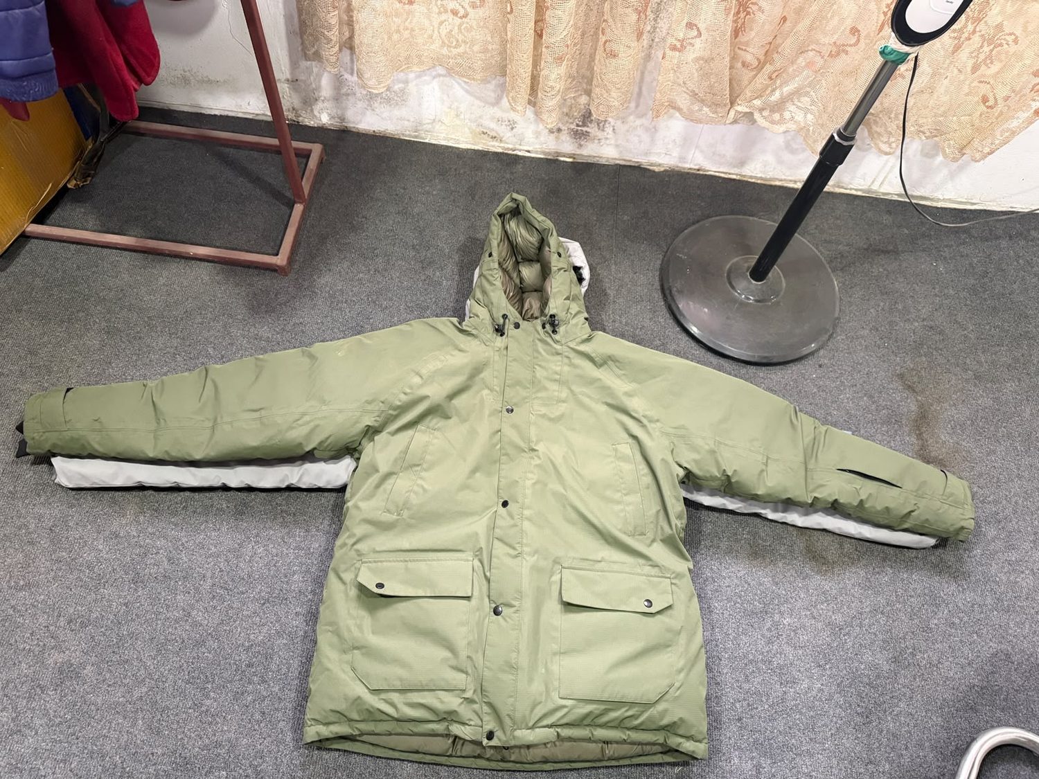 An olive green hooded parka laid flat showing snap studs and flapped lower pockets