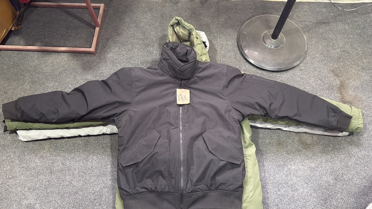 A charcoal jacket laid over an olive one on grey carpet with a handwritten paper tag at the front