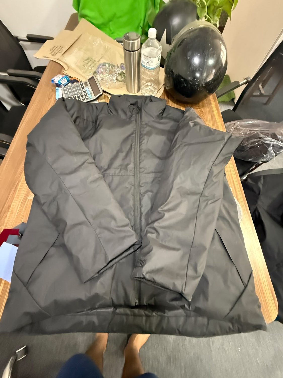 A dark grey jacket lying open on a wooden table with a calculator, a flask and a black helmet along the far edge